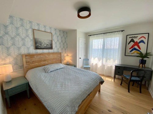 Bright bedroom featuring a wooden bed, patterned wallpaper, a desk, and large windows with sheer curtains.