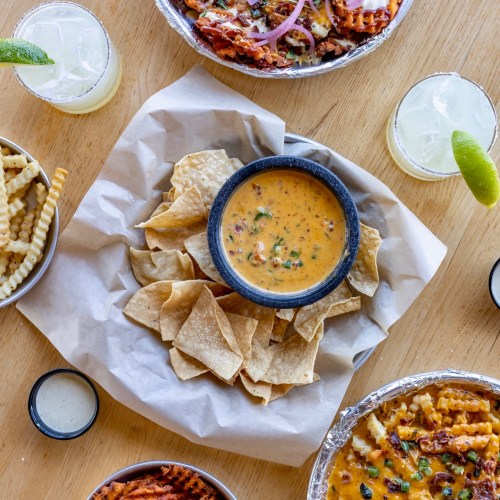 A spread of delicious food featuring tortilla chips with cheese dip, fries, and two bowls of savory dishes on a wooden table.