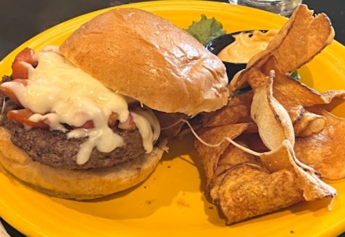 A juicy burger topped with melted cheese and tomatoes, served with crispy potato chips on a yellow plate.