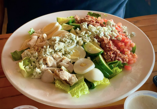 A colorful salad with greens, chicken, hard-boiled eggs, tomatoes, and crumbled cheese on a white plate.