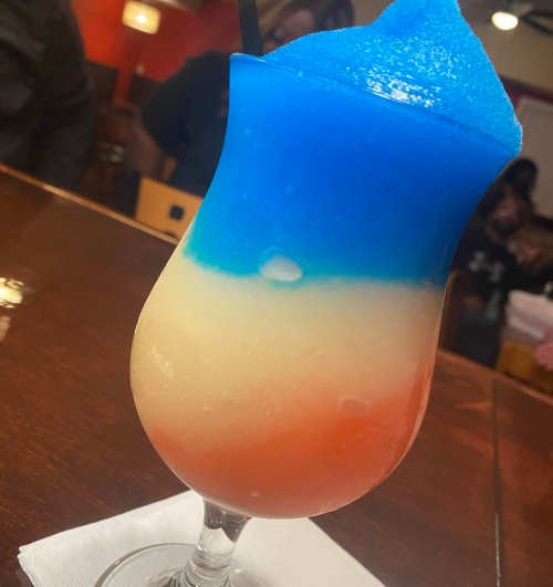 A colorful layered drink with blue, white, and red slushy textures in a tall glass, sitting on a wooden table.