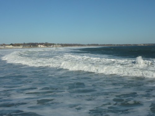 Waves gently roll onto a sandy beach under a clear blue sky, with distant shoreline visible in the background.