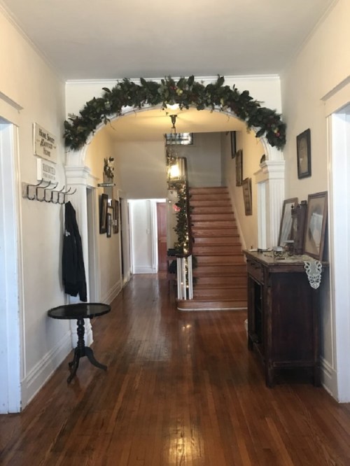 A hallway with wooden floors, a staircase, and festive decorations above an archway. Walls adorned with framed pictures.