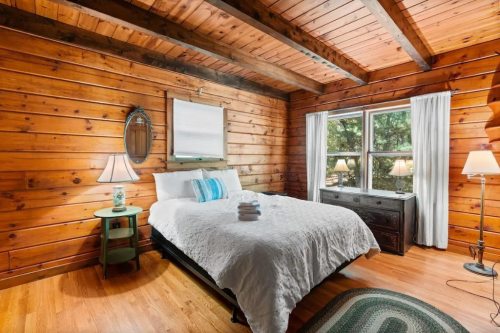 Cozy wooden bedroom with a bed, nightstands, and large windows, featuring warm tones and rustic decor.