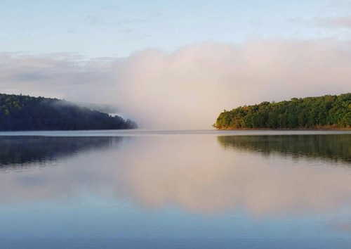 A serene lake surrounded by lush green hills, with mist rising gently from the water under a soft, cloudy sky.