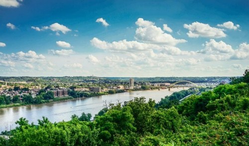 A scenic view of a river surrounded by greenery, with a city skyline and blue sky filled with fluffy clouds.