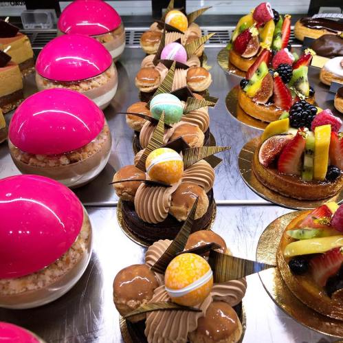 A display of colorful desserts, including glossy pink mousse, chocolate pastries, and fruit-topped tarts.