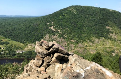 A rocky outcrop overlooks a lush green hillside and a winding river below, under a clear blue sky.
