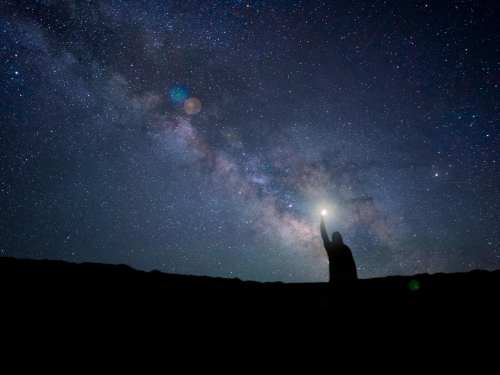 Silhouette of a person reaching towards a starry night sky filled with the Milky Way.