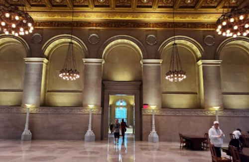 Elegant interior of a grand hall with arched windows, chandeliers, and decorative ceilings, featuring people and furniture.