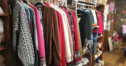 A variety of colorful sweaters hanging on a rack in a cozy shop, with shelves of items in the background.