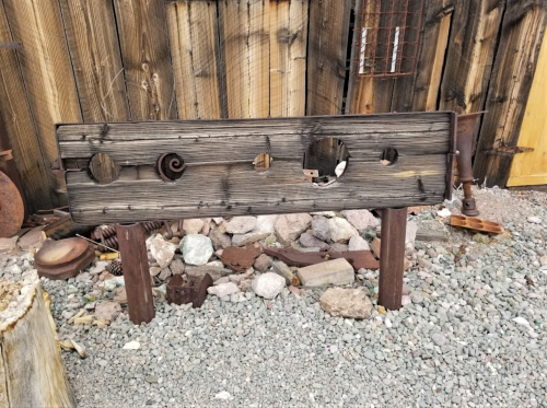 A weathered wooden sign with circular cutouts, set against a rustic wooden backdrop and surrounded by gravel and debris.