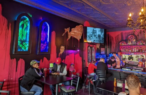 A vibrant bar interior with colorful stained glass, brick accents, and patrons enjoying drinks at tables.