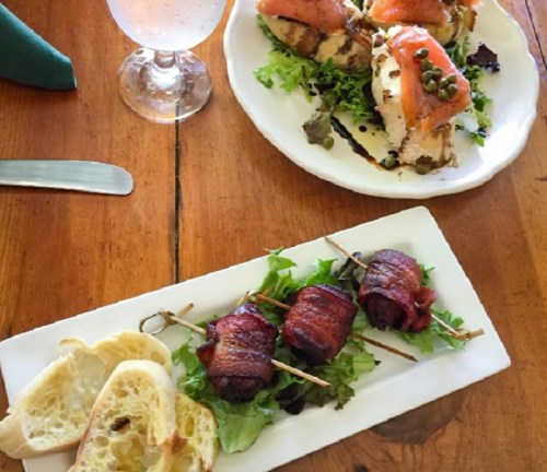 A plate of bacon-wrapped meat on greens, accompanied by bread, with a second dish of salmon on toast in the background.