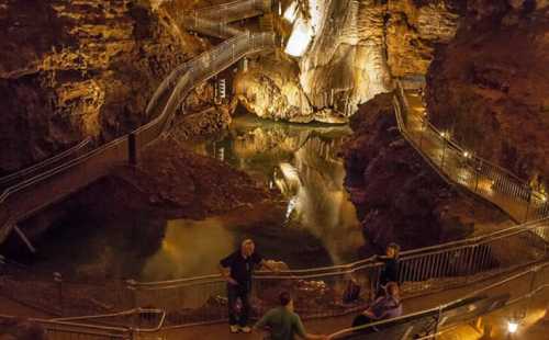 A dimly lit cave with winding walkways, a reflective pool, and visitors exploring the natural rock formations.