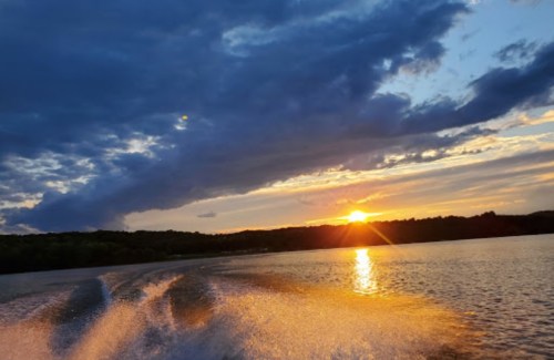 A sunset over a lake, with vibrant clouds and water trails from a boat in the foreground.
