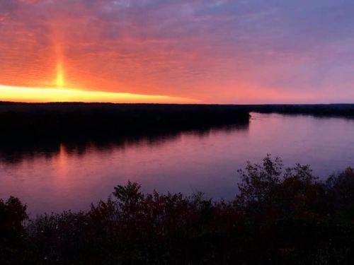 A serene river at sunset, reflecting vibrant orange and purple hues in the sky and water, surrounded by trees.