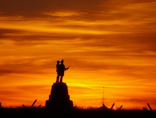 Silhouette of a statue depicting two figures against a vibrant orange and yellow sunset sky.