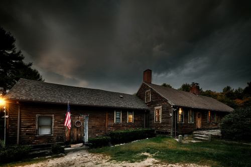 A rustic wooden house under a dark, cloudy sky, featuring an American flag and warm lights glowing from the windows.