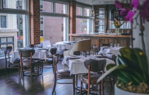 Elegant restaurant interior with wooden floors, white tablecloths, and large windows showcasing a city view.