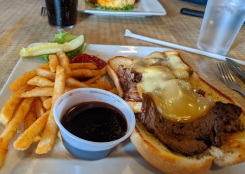 A plate with a beef sandwich topped with melted cheese, fries, a pickle, and a small cup of sauce.