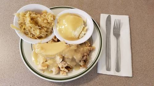 A plate of chicken with gravy, served with mashed potatoes and sauerkraut, alongside a knife and fork on a napkin.