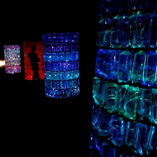 Colorful illuminated structures made of bottles, with a silhouette of a person in the background, set against a dark space.