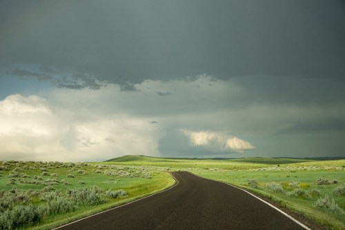 A winding road stretches through lush green fields under a dramatic sky with dark clouds.