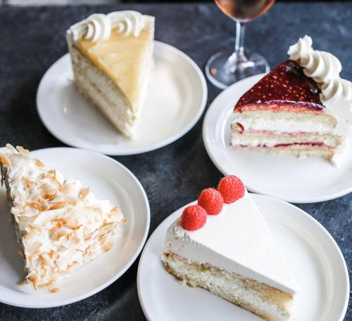 Four slices of cake on white plates: lemon, raspberry, coconut, and strawberry, with a glass of rosé in the background.