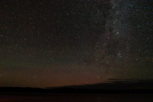 A starry night sky filled with countless stars over a calm body of water, with a hint of the Milky Way visible.