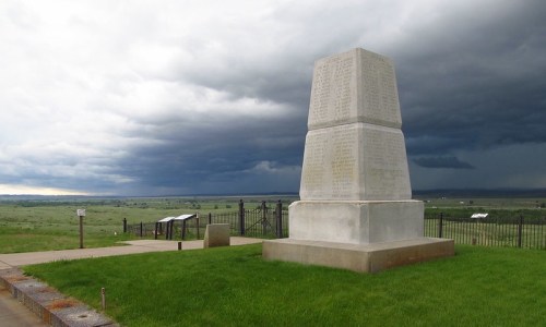 A tall stone monument stands on green grass, with dark storm clouds looming in the background over a vast landscape.
