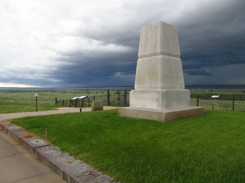 A tall monument stands on green grass, with dark storm clouds looming in the background over a vast landscape.
