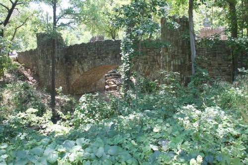A stone bridge arches over a creek, surrounded by lush greenery and trees in a serene natural setting.