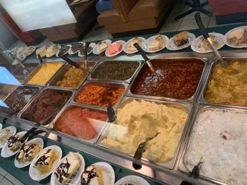 A buffet display featuring various dishes, including meats, vegetables, mashed potatoes, and desserts in plastic plates.