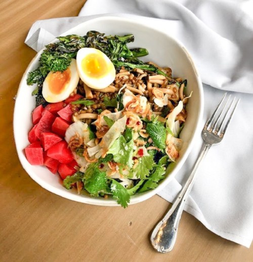 A bowl of colorful salad with greens, watermelon, soft-boiled egg, and noodles, served with a fork on a wooden table.