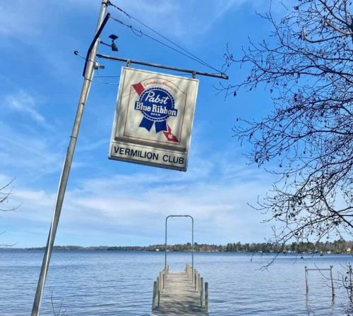A wooden dock extends into a calm lake, with a sign for the Vermilion Club hanging above. Clear blue sky in the background.