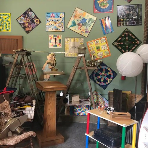 A vintage shop interior featuring various items, including games, furniture, and decor, against a green wall.