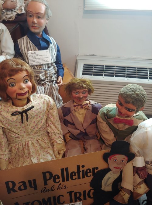A collection of vintage ventriloquist dolls, featuring various expressions and outfits, displayed with a sign for Ray Pelletier.