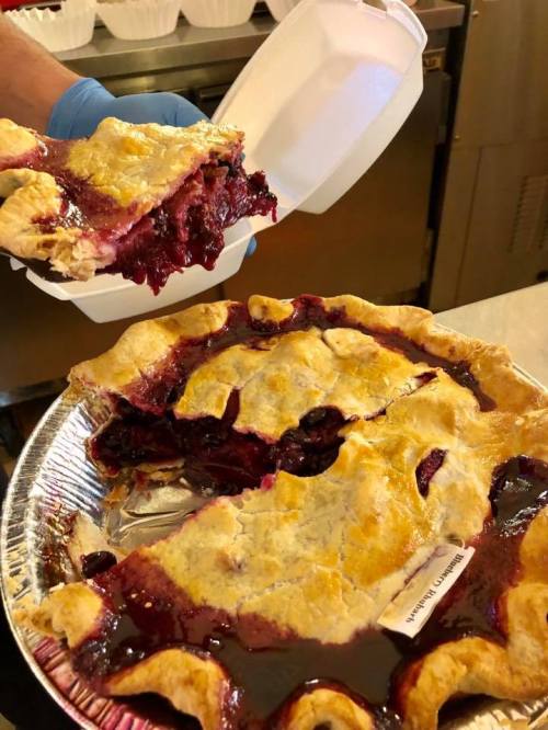 A hand holds a takeout container above a freshly baked cherry pie with a golden crust and juicy filling.