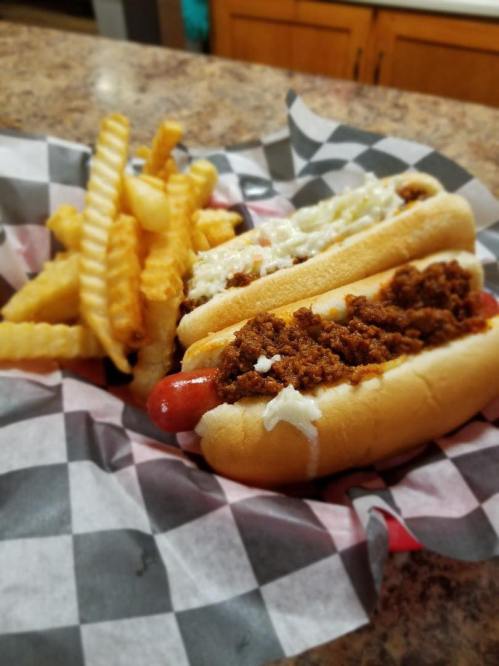 Two hot dogs topped with chili and mayonnaise, served with a side of crinkle-cut fries in a checkered paper basket.