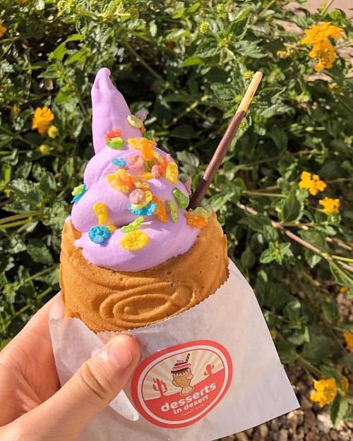 A hand holding a purple soft serve ice cream cone topped with colorful sprinkles, set against a backdrop of green foliage and flowers.
