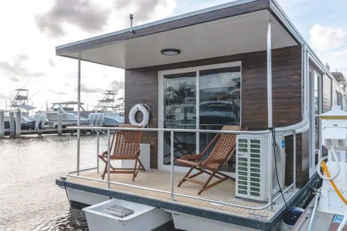 A modern houseboat with two chairs on the deck, docked at a marina with boats in the background.