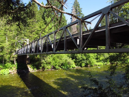 A metal bridge spans a clear river, surrounded by lush green trees and sunlight filtering through the foliage.