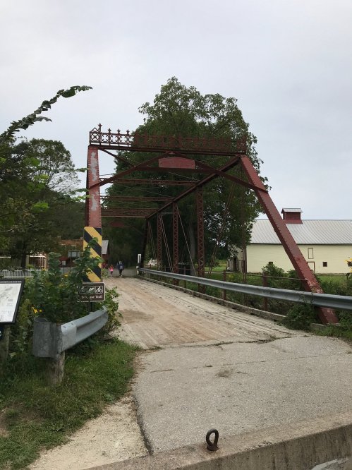 A red metal bridge spans a dirt road, surrounded by trees and a building in the background under a cloudy sky.