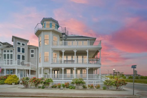 A large, multi-level beach house with a wraparound porch, surrounded by flowers, against a colorful sunset sky.