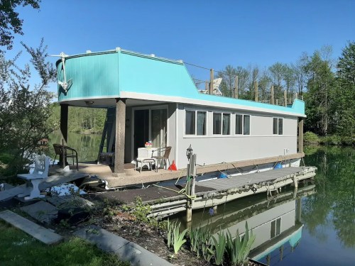 A colorful houseboat on a calm river, surrounded by greenery and featuring a small deck with outdoor furniture.