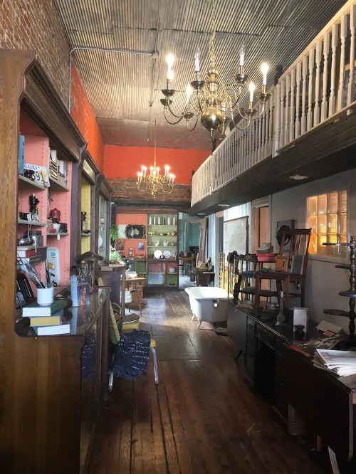 A cozy, vintage shop interior with wooden floors, chandeliers, and various furniture pieces displayed along the walls.