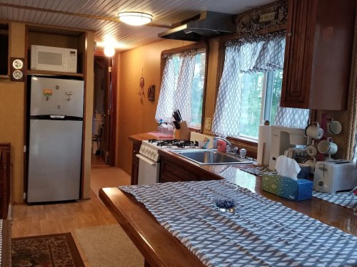 A cozy kitchen with a wooden table, refrigerator, stove, and large windows, featuring light-colored decor and a checkered tablecloth.
