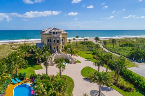 Aerial view of a beachfront mansion with a pool, surrounded by palm trees and lush greenery, overlooking the ocean.