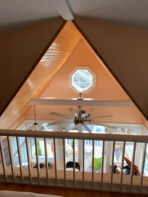 A cozy attic space with a triangular wooden ceiling, a ceiling fan, and a view of a bright room below.